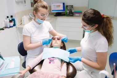 Ofiste destal sandalyede oturan 3d gözlükküçük kız ve yıllık kontrol var. Asistanlı diş hekimi