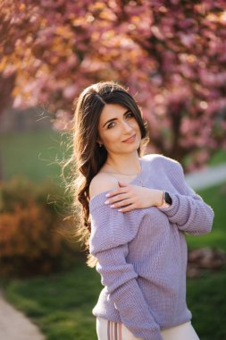 Parktaki sakura ağacının önünde mor bir kazak standında çekici genç kadın