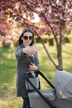 Genç anne bebeğiyle parkta yürüyor ve bir selfie çekiyor.