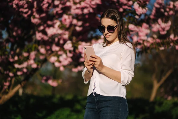 Jonge vrouw in wit shirt gebruik smartphone buiten. Achtergrond van de roze boom — Stockfoto