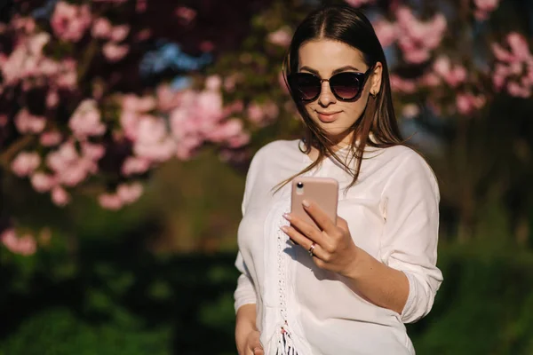 Jonge vrouw in wit shirt gebruik smartphone buiten. Achtergrond van de roze boom — Stockfoto