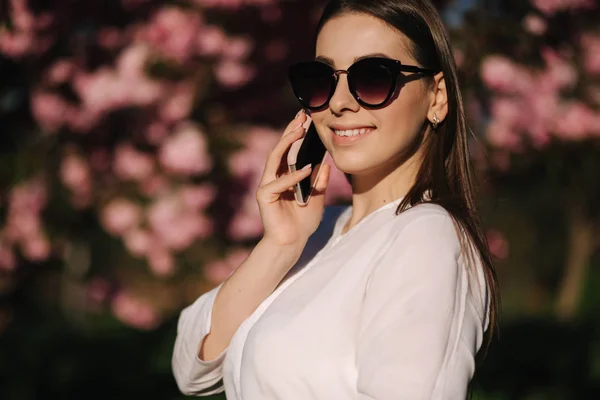 Retrato de mulher jovem attravtive em camisa branca e óculos de sol usar smartphone fora. Fala com alguém ao pé do telefone. Fundo da árvore rosa — Fotografia de Stock