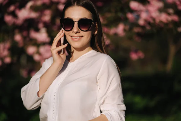 白いシャツとサングラスをかけた若い女性の肖像画は、外でスマートフォンを使用しています。電話で何とかして皮をむく。ピンクの木の背景 — ストック写真