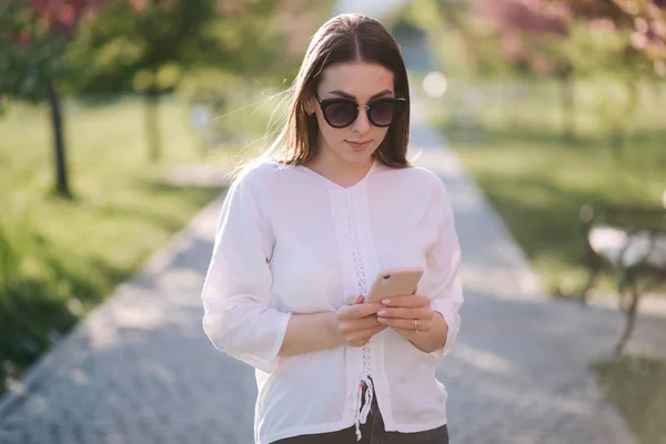 Jonge vrouw in wit shirt gebruik smartphone buiten. Achtergrond van de roze boom — Stockfoto