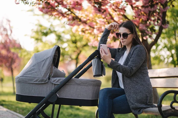 若いお母さんはベビーカーで赤ちゃんとベンチに座って電話を使います。インターネットで何かを見て — ストック写真
