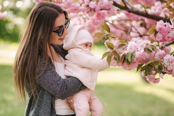 Mother show firs time to her daughter pink tree. Happy baby hold branch of sakura. Side view — Stock Photo, Image