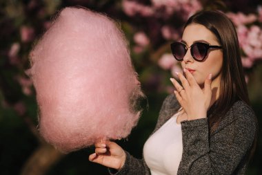 Elinde pamuk şeker tutan gülümseyen kızın yakın portresi. Grrey blazer ve güneş gözlüğü giymiş kız. Pamuk şeker yiyen kız