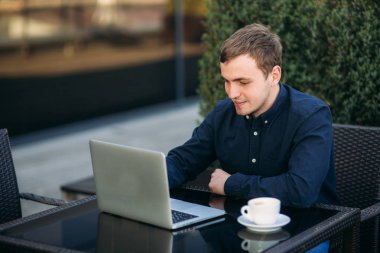 Genç adam laptopuyla ve işiyle kafede oturuyor. Fedai haberleri var. Cappuchino masada.