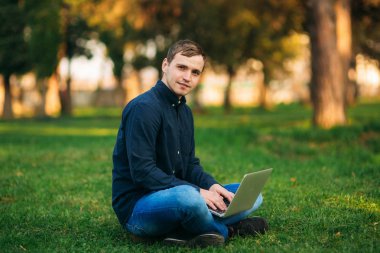 Parktaki çimlerde oturan ve dizüstü bilgisayarda çalışan genç adam.