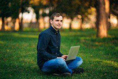 Parktaki çimlerde oturan ve dizüstü bilgisayarda çalışan genç adam.