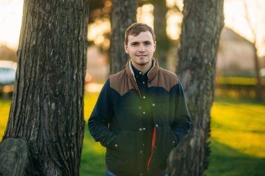 Koyu mavi gömlekli genç adam parkta fotoğrafçıya poz veriyor.