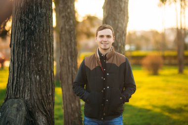 Koyu mavi gömlekli genç adam parkta fotoğrafçıya poz veriyor.