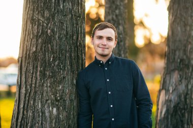 Koyu mavi gömlekli genç adam parkta fotoğrafçıya poz veriyor.