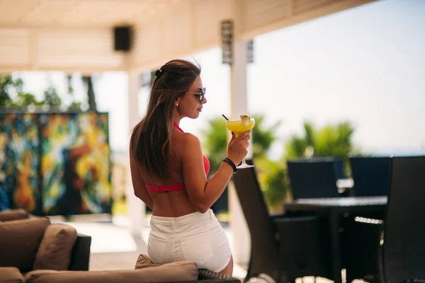 Gyönyörű fiatal nő visel fürdőruhát, ivás, egy színes koktél ül egy légiutas-kísérő, a beach club bárban. Lenyűgöző lány neki italt a nyári idő — Stock Fotó