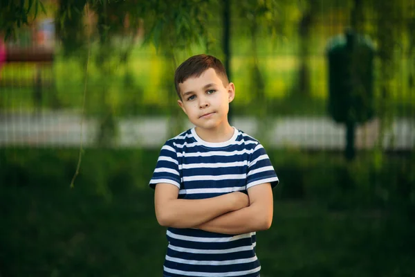 En liten pojke i en randig T-shirt står framför grön bakgrund. Leende och ser att fotografen — Stockfoto