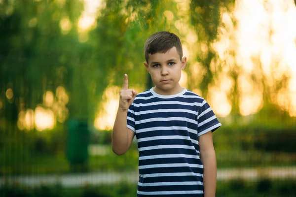 En liten pojke i en randig T-shirt stående framför grön bakgrund. Leende och ser till fotografen. Pojken satte fingret upp — Stockfoto