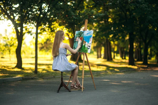 Krásné blond vlasy dívka sedí na stoličce a nakreslí obrázek v parku pomocí palety barvy a stěrky — Stock fotografie