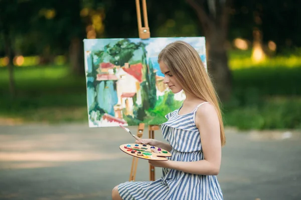 Красива світле волосся дівчина сидить на табуреті і малює фотографію в парку, використовуючи палітру з фарбами і шпателем — стокове фото