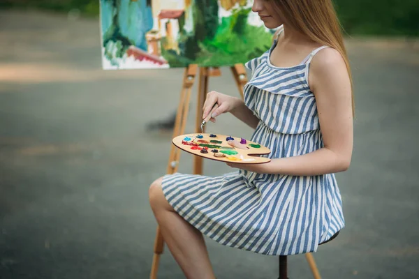 Piękne blond włosy dziewczyna siedzi na stołku i rysuje obraz w parku przy użyciu palety farb i łopatki — Zdjęcie stockowe