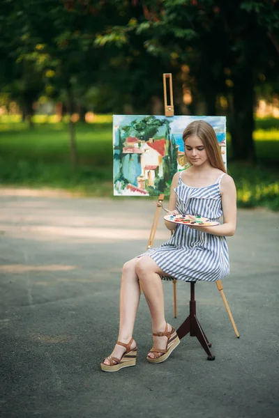 Hermosa chica de pelo rubio sentada en el taburete y dibuja un cuadro en el parque usando una paleta con pinturas y una espátula — Foto de Stock