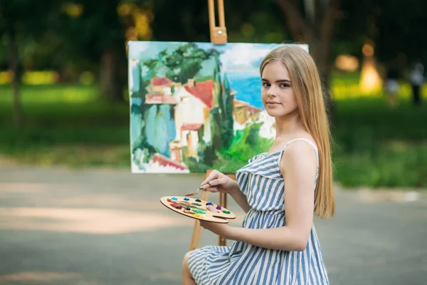 Hermosa chica de pelo rubio sentada en el taburete y dibuja un cuadro en el parque usando una paleta con pinturas y una espátula —  Fotos de Stock