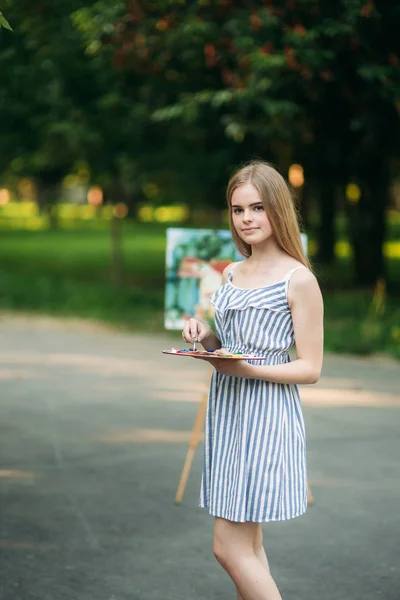 Beautiful girl draws a picture in the park using a palette with paints and a spatula. Easel and canvas with a picture. Background of picture