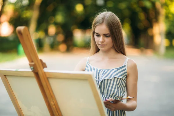 Menina bonita desenha um quadro no parque usando uma paleta com tintas e uma espátula. Easel e lona com uma imagem — Fotografia de Stock