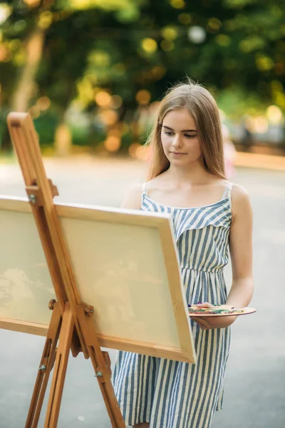 Piękna dziewczyna rysuje obraz w parku przy użyciu palety farb i łopatką. Sztalugi i płótno z rysunkiem — Zdjęcie stockowe