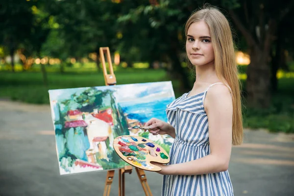 Beautiful girl artist draws a picture in the park using a palette with paints and a spatula. Easel and canvas with a picture