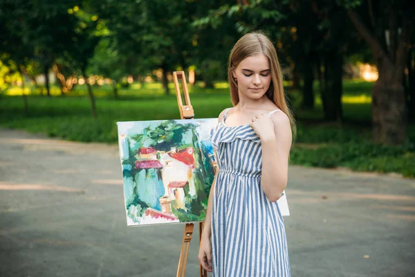 Belle fille artiste dessine une image dans le parc en utilisant une palette avec des peintures et une spatule. Chevalet et toile avec une image — Photo