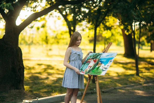Mooi meisje kunstenaar tekent een afbeelding in het park met behulp van een palet met verf en een spatel. Ezel en canvas met een afbeelding — Stockfoto