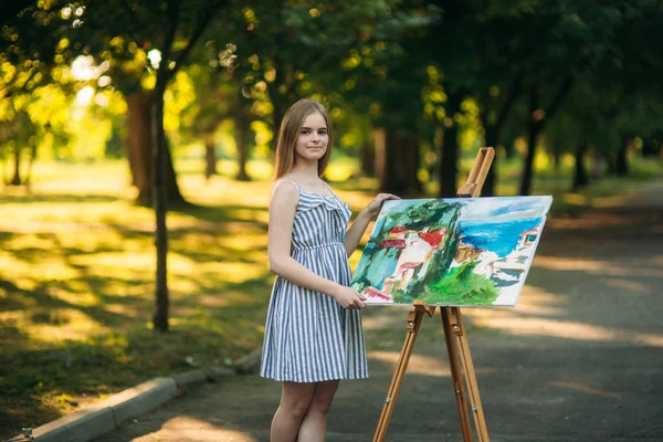 Krásná dívka umělec nakreslí obrázek v parku pomocí palety barvy a stěrky. Malířský stojan a plátno s obrázkem — Stock fotografie