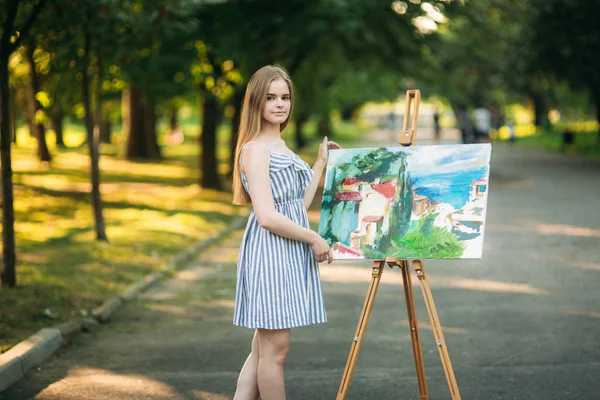 Beautiful girl artist draws a picture in the park using a palette with paints and a spatula. Easel and canvas with a picture