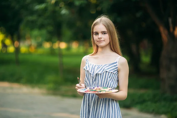 Hermosa artista chica dibuja un cuadro en el parque utilizando una paleta con pinturas y una espátula. caballete y lienzo con una imagen —  Fotos de Stock