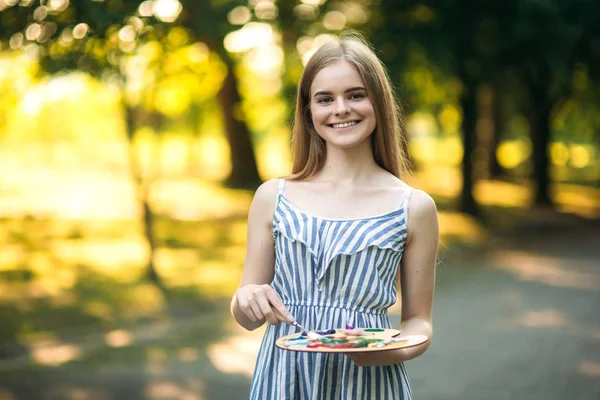 Hermosa artista chica dibuja un cuadro en el parque utilizando una paleta con pinturas y una espátula. caballete y lienzo con una imagen — Foto de Stock