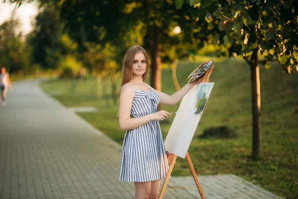 Schöne Künstlerin steht im Park vor dem Bild und hält Palette mit Farben — Stockfoto
