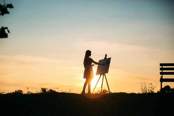 Silhueta de uma menina loira pinta uma pintura na tela com a ajuda de tintas. Um cavalete de madeira mantém a imagem. O verão é um dia ensolarado, pôr do sol — Fotografia de Stock