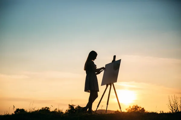 Silhouette of a blonde girl paints a painting on the canvas with the help of paints. A wooden easel keeps the picture. Summer is a sunny day, sunset