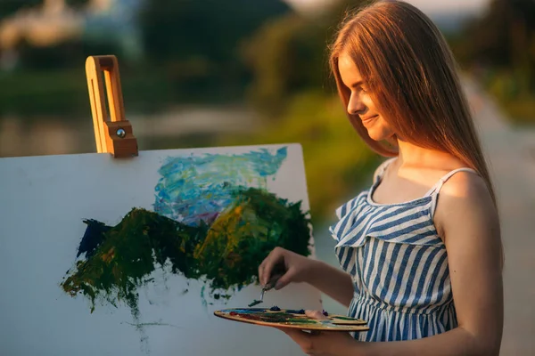 Retrato de bela menina de cabelo loiro desenha quadro pelo rio — Fotografia de Stock