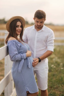 Mutlu bir aile bebek bekliyor. Hamile kadın ve kocası dışarıda çiftliğin yanında duruyorlar. Mavi elbiseli kadın ve beyaz erkek