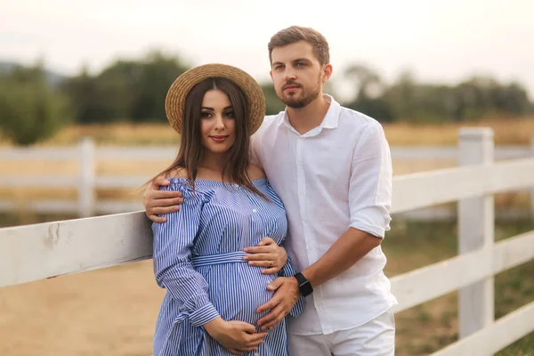 La familia feliz está esperando al bebé. La mujer embarazada y su esposo afuera están junto a la granja. Mujer en vestido azul y hombre en blanco —  Fotos de Stock