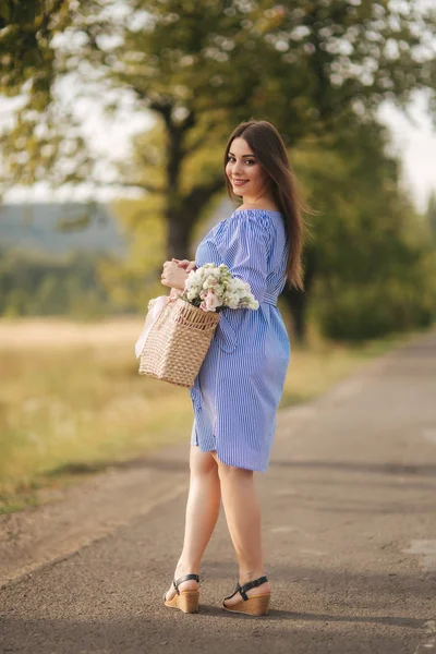 Schöne schwangere Frau in blauem Kleid, die den Hof betritt. Stricktasche mit Blumenstrauß. Hintergrund von Baum und Bergen — Stockfoto