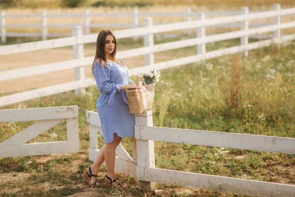 Mulher grávida bonita em vestido azul andando ouvir a fazenda. Saco de malha com buquê de flores. Fundo de árvore e montanhas — Fotografia de Stock