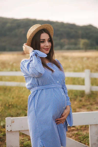 La donna incinta in natura prova il cappello lavorato a maglia. Sfondo di campo e recinto bianco vicino alla fattoria — Foto Stock