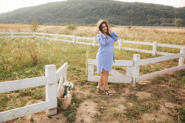 Femme enceinte dans la nature essayer chapeau tricoté. Fond de champ et clôture blanche près de la ferme — Photo