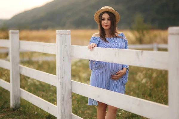 Belle femme enceinte en robe bleue et chapeau tricoté debout près de la clôture dans la ferme. Elle mit les mains sur son ventre et sourit — Photo