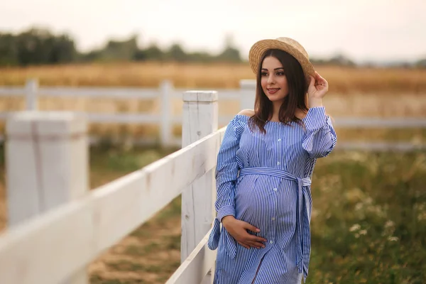 Mulher grávida bonita em vestido azul e chapéu de malha ficar perto da fance na fazenda. Ela colocou as mãos na barriga e sorriu — Fotografia de Stock