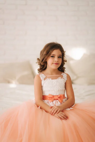 Little beautiful girl with brown hair in a Peach-colored dress. Girl sitting on the sofa