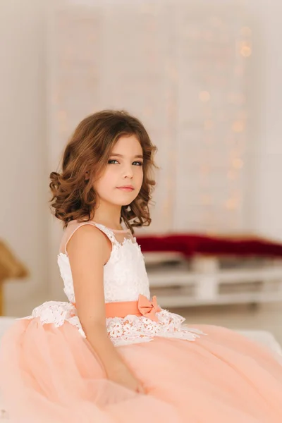 Menina bonita pequena com cabelo castanho em um vestido de cor de pêssego. Menina sentada no sofá — Fotografia de Stock