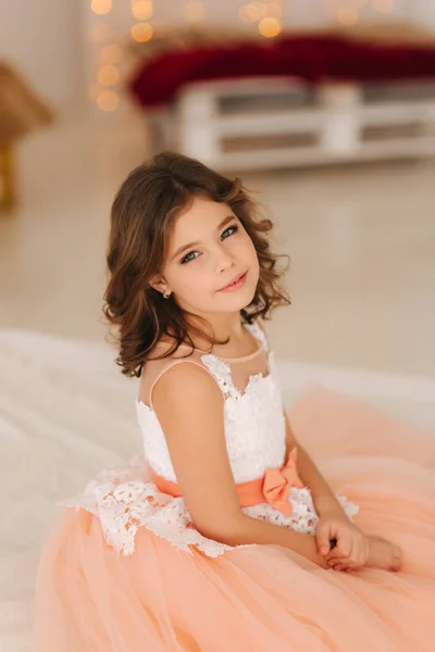 Little beautiful girl with brown hair in a Peach-colored dress. Girl sitting on the sofa — Stock Photo, Image